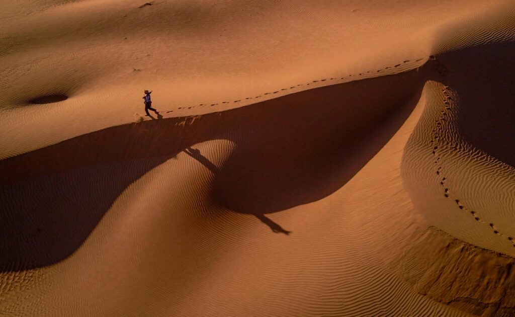 A lone person walks across sand dunes at sunset, casting long shadows in the desert.